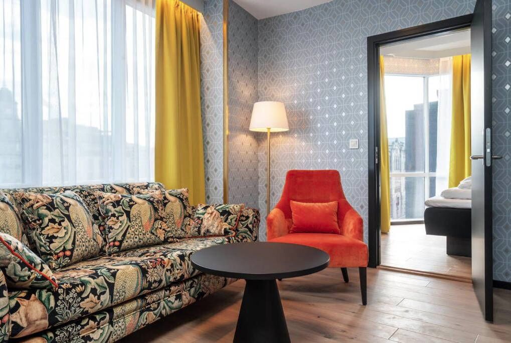 A room with a patterned sofa, a red armchair, a small round table, a lamp, and a view through a door.