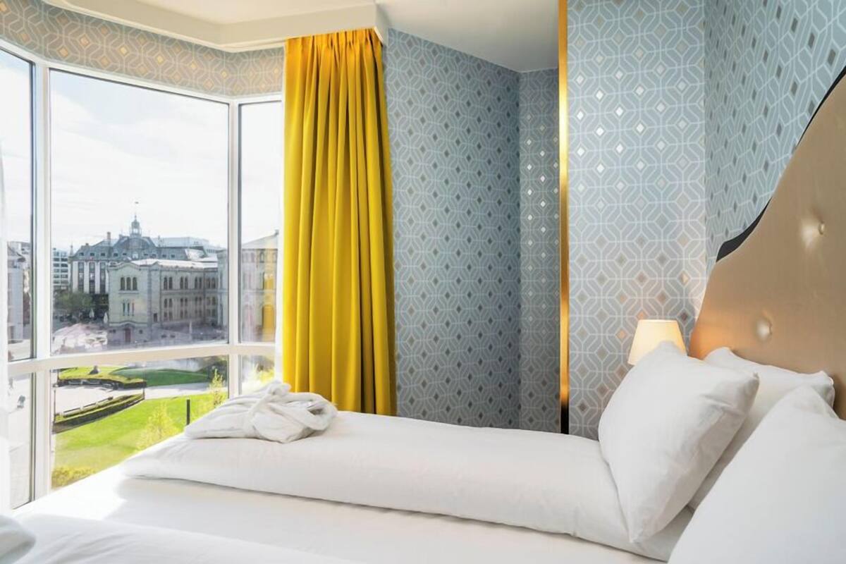 A hotel room with a large window, a bed with white linens, a yellow curtain, patterned wallpaper, and a nightstand with a lamp.