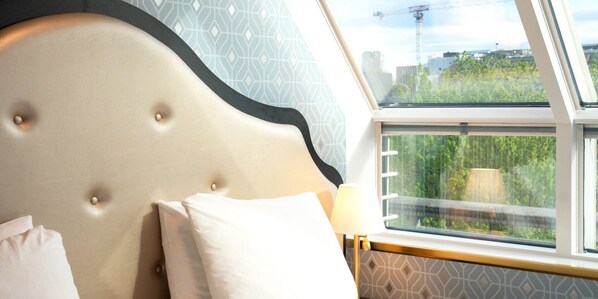 A hotel room with a tufted headboard, a large window with a view of greenery and buildings, and a patterned wall.
