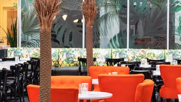 Ein Restaurant mit orangefarbenen Stühlen und Tischen, großen Fenstern mit tropischen Blattmustern und Palmen.