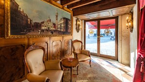 A room with a large framed painting, wooden paneling, two upholstered chairs, a small round table, and a view through a glass door to an outdoor area with a red awning. - Hotel Scandinavia - Relais (Venice)