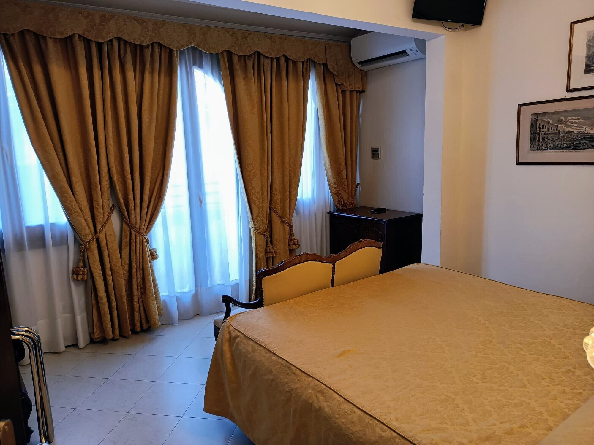 A hotel room with a bed, a desk, and a chair. There is a window with curtains, an air conditioning unit, and a framed picture on the wall.