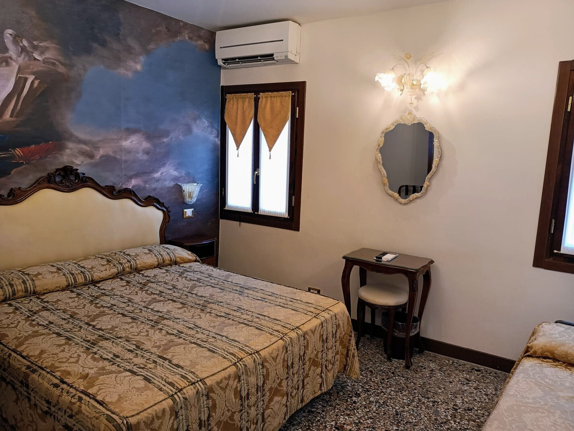 Quarto com cama grande, mesa pequena com cadeira, espelho e janela com cortinas.