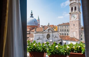 A view of historic buildings with a prominent clock tower and a dome, viewed through a window with potted plants in the foreground.