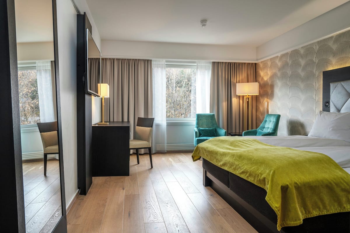 A hotel room with a bed, two chairs, a desk, and a large window.