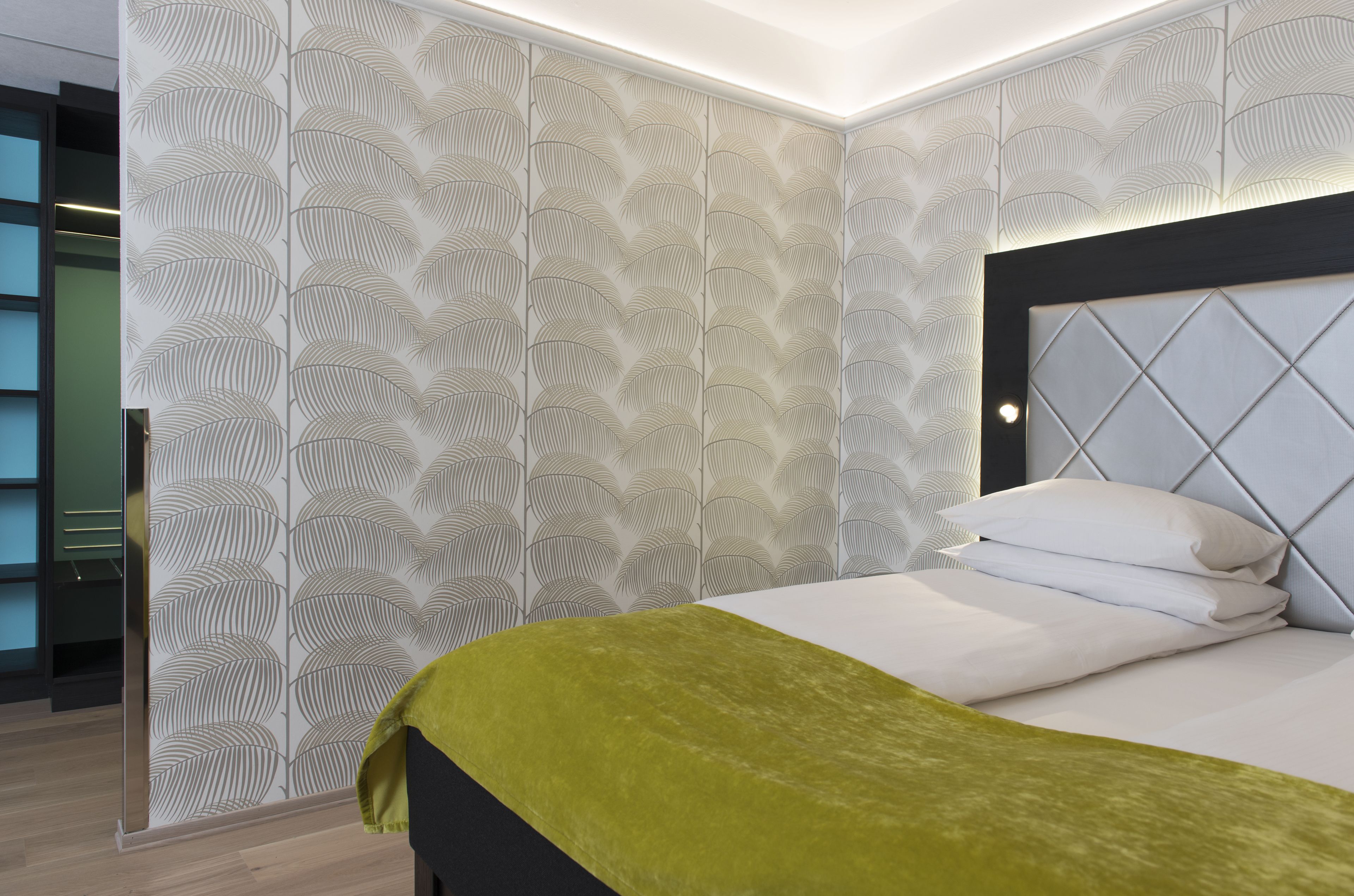 Habitación de hotel moderna con papel tapiz estampado, cabecero capitonado y una cama bien hecha con sábanas blancas y una manta verde.