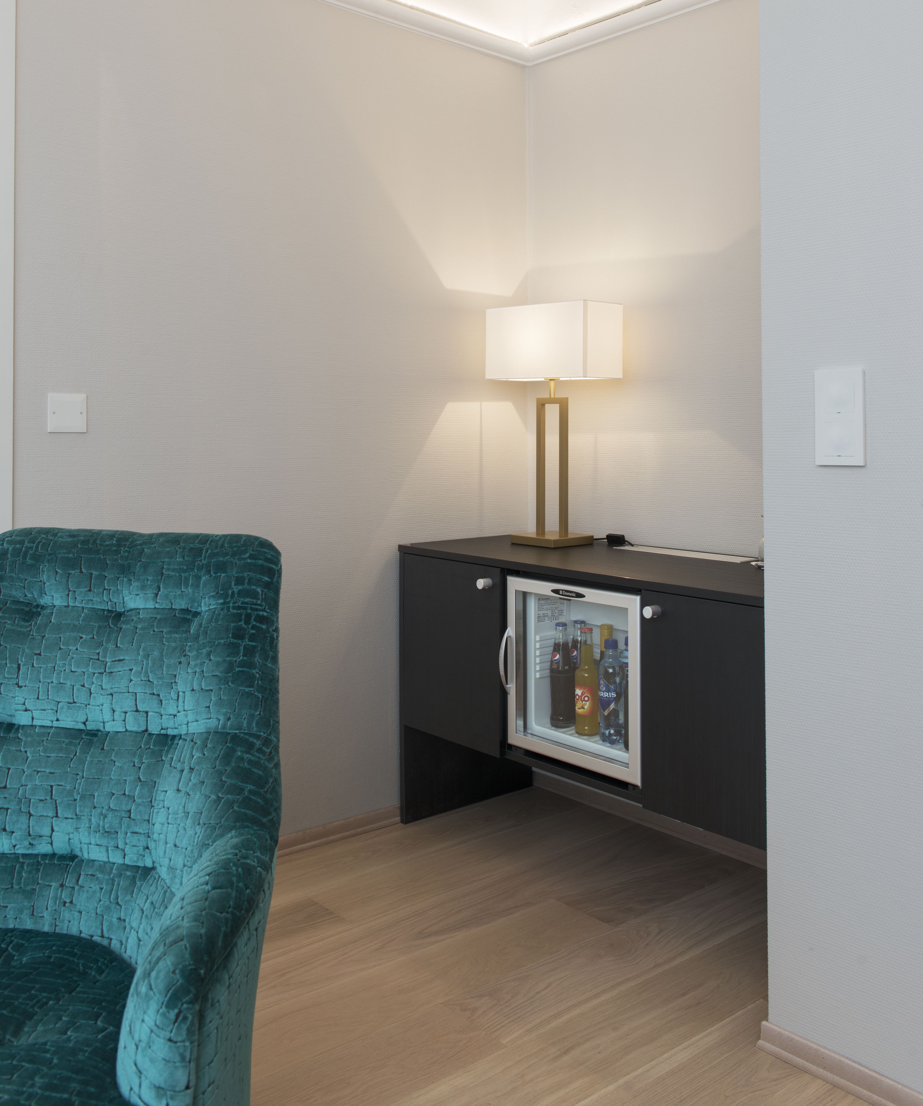 A corner room with a teal chair, a minibar, and a wall-mounted lamp.