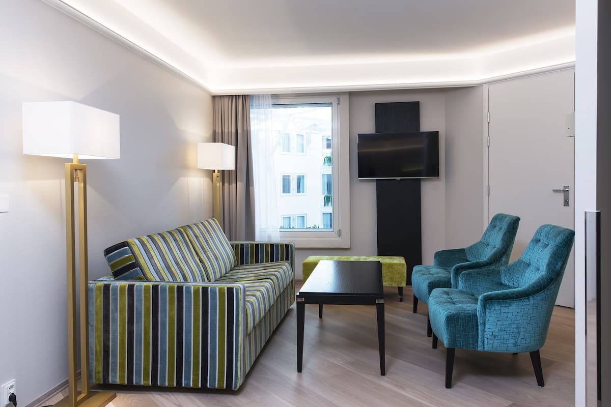 A modern living room with a striped sofa, a small table, two armchairs, a floor lamp, a TV mounted on the wall, and a window with a view of buildings.