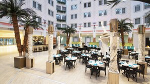 Un atrium d’hôtel comprenant de nombreuses tables et chaises, des palmiers et un espace bar.