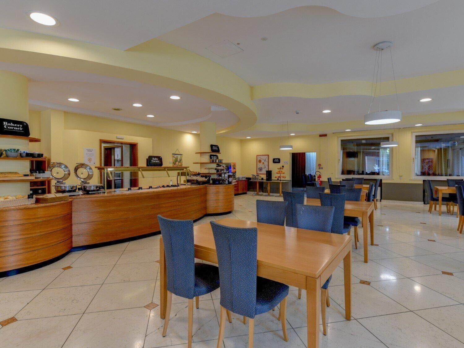 A spacious dining area with multiple tables and chairs, a central counter with various food items, and a clean, well-lit environment.