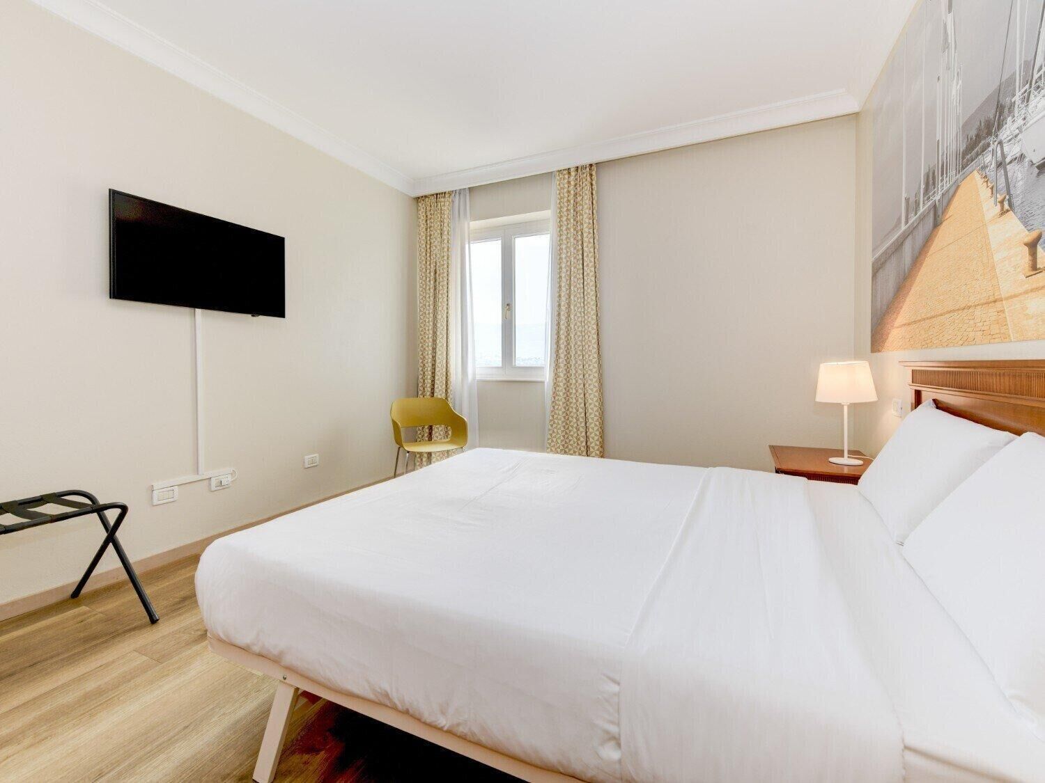 Ein Hotelzimmer mit einem Bett, einem an der Wand befestigten Fernseher, einem Sessel, einem Fenster mit Vorhängen und einer Lampe auf einem Nachttisch.