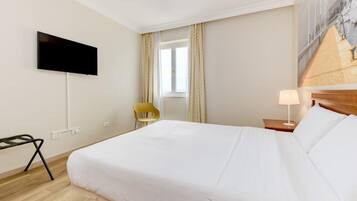 Ein Hotelzimmer mit einem Bett, einem an der Wand befestigten Fernseher, einem Sessel, einem Fenster mit Vorhängen und einer Lampe auf einem Nachttisch.
