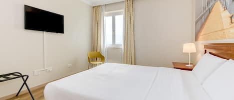 A hotel room with a bed, a TV mounted on the wall, a chair, a window with curtains, and a lamp on a bedside table.
