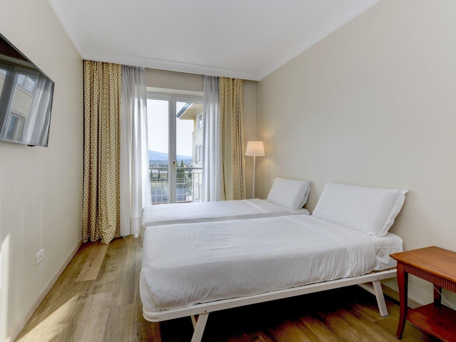 A hotel room with two beds, a bedside table, a lamp, and a window with curtains.