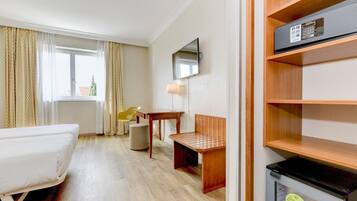 A hotel room with a bed, a desk, a chair, a TV, and a minibar.