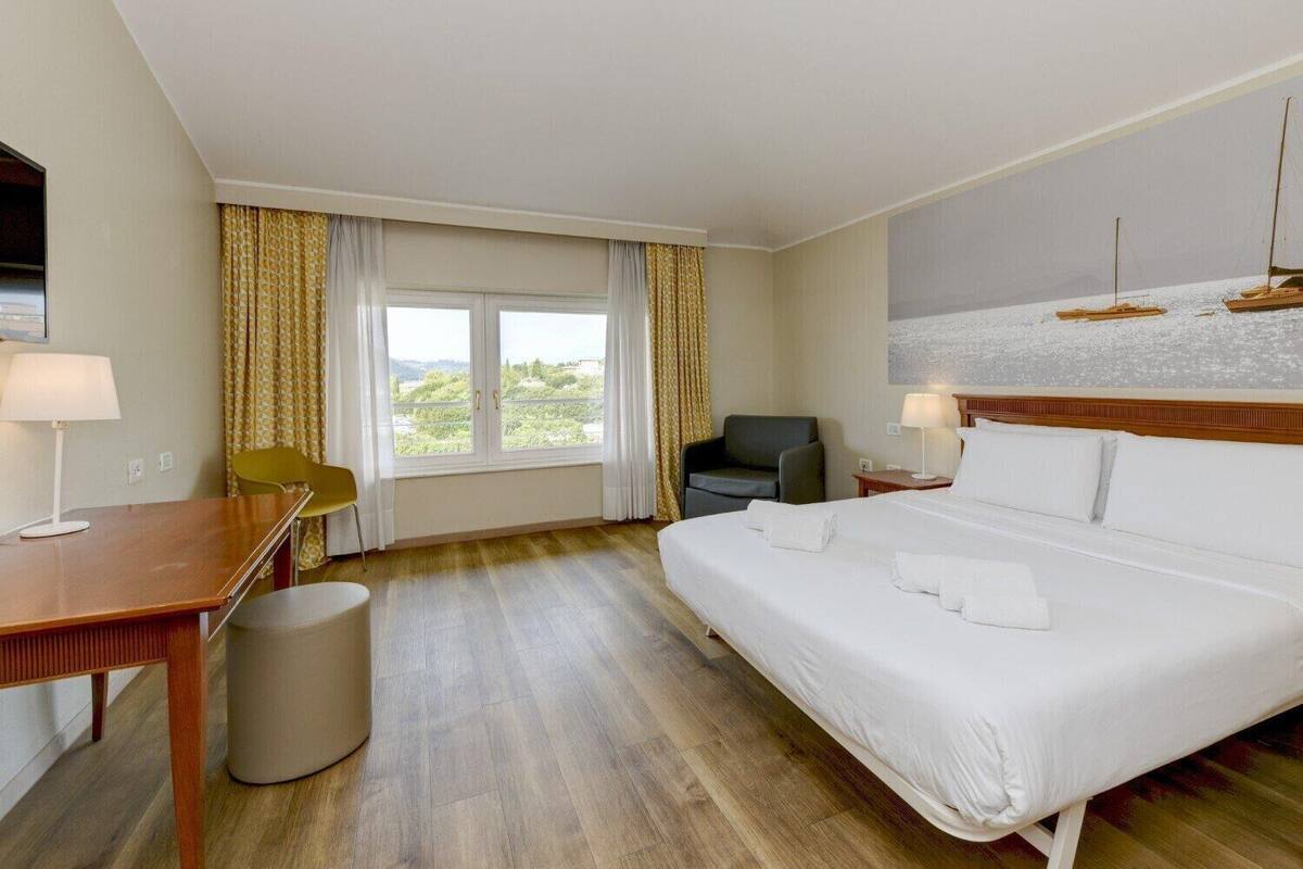 Une chambre d’hôtel avec un grand lit, un bureau avec une chaise, une petite poubelle et une fenêtre avec des rideaux.