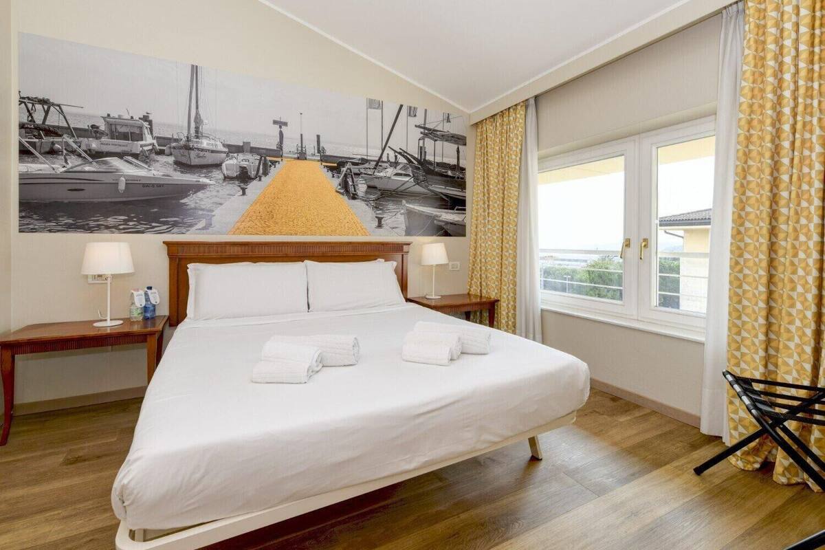 Une chambre d’hôtel avec un grand lit, une tête de lit en bois, deux tables de chevet avec des lampes, une grande fenêtre avec des rideaux et une photographie en noir et blanc accrochée au mur.