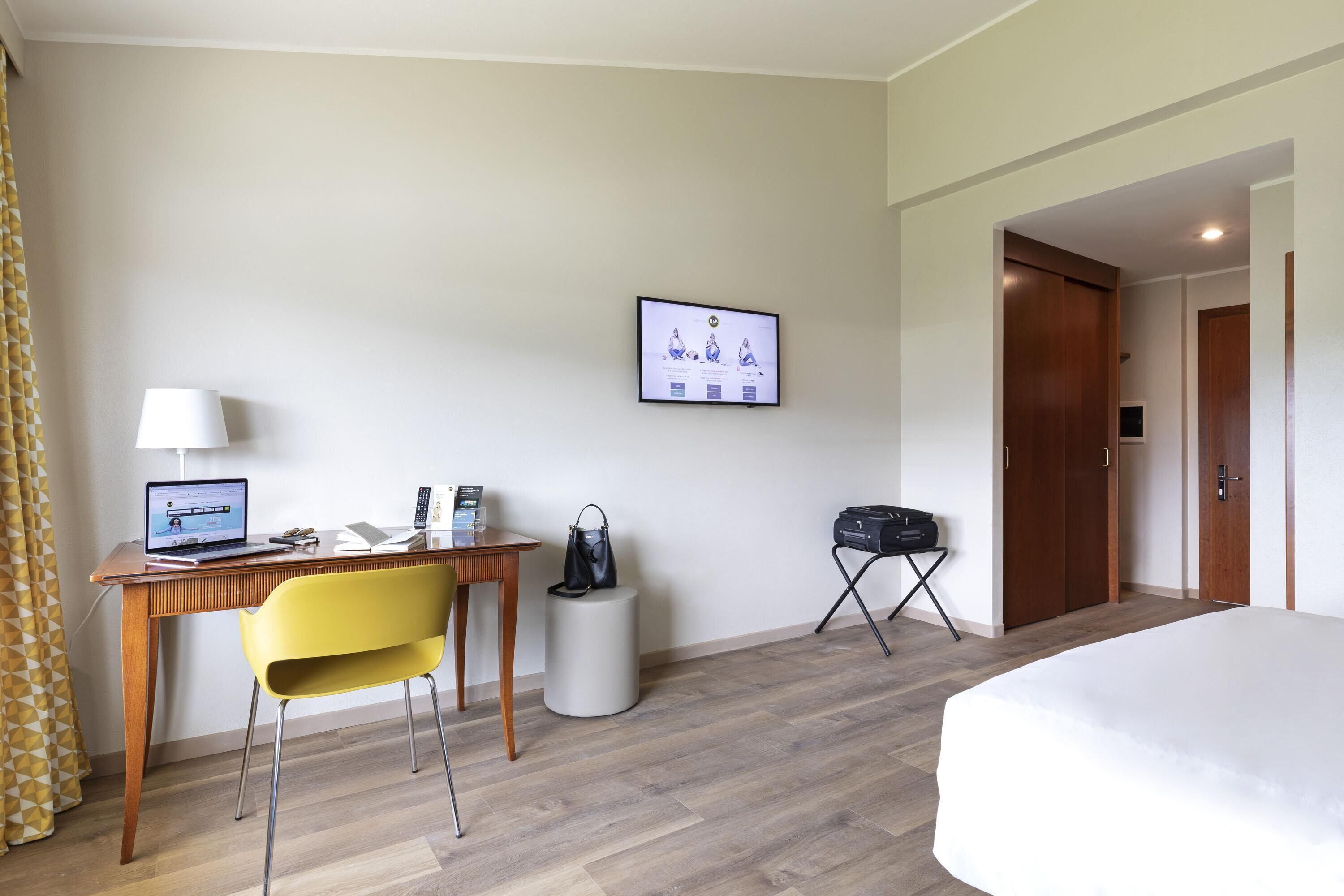 Habitación de hotel con cama, escritorio con computadora portátil, silla, mesita, cesto de basura y perchero.