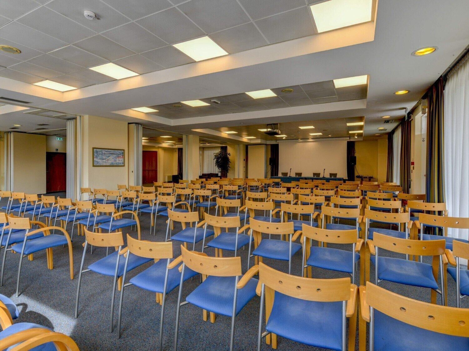 A conference room with rows of wooden chairs and blue cushions, equipped with ceiling lights and a projector screen.