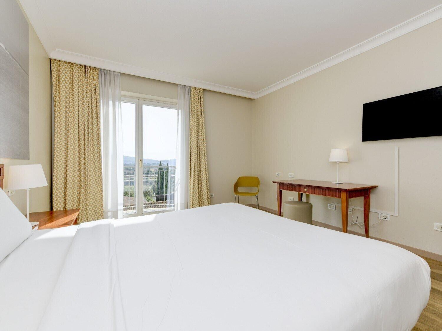 A hotel room with a large bed, a desk, a chair, and a TV on the wall.