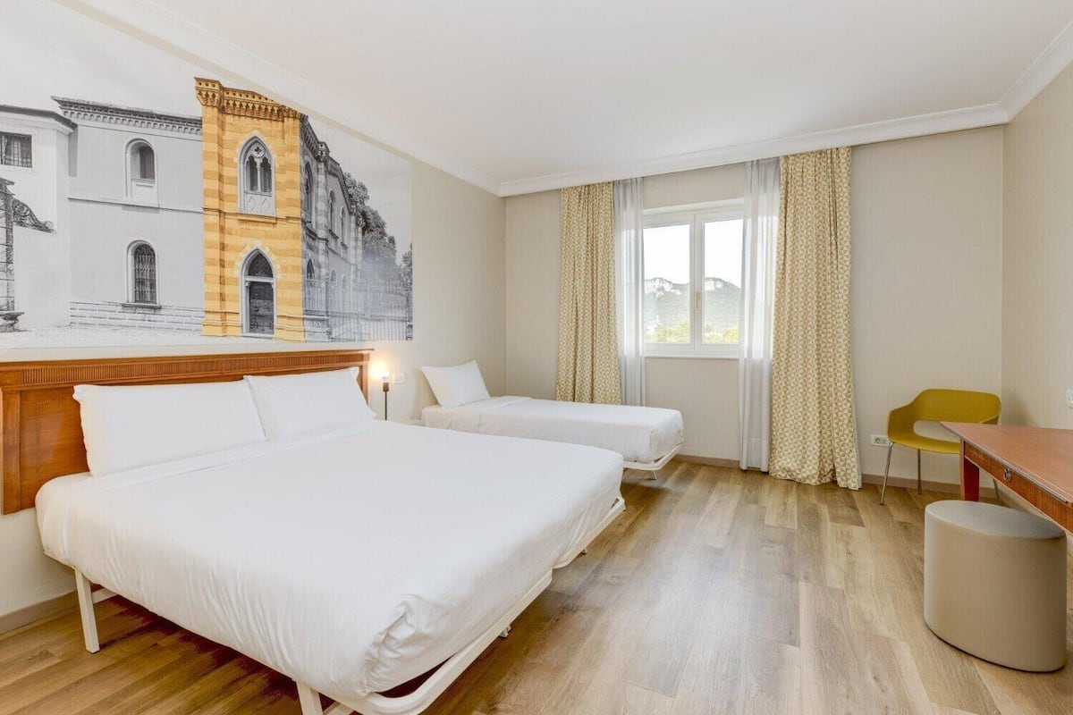 Une chambre d’hôtel avec deux lits, une tête de lit en bois, un bureau, une chaise et une vue sur un immeuble par la fenêtre.