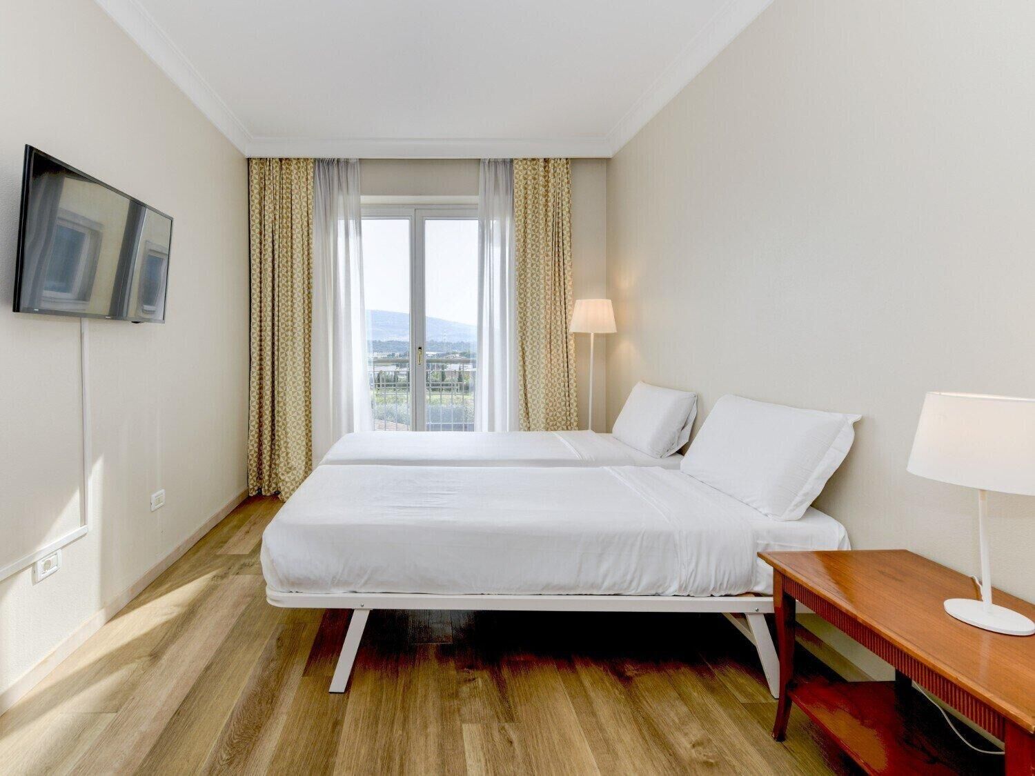 Une chambre d’hôtel avec un lit, une table de chevet, une lampe, un téléviseur fixé au mur et une fenêtre avec des rideaux.