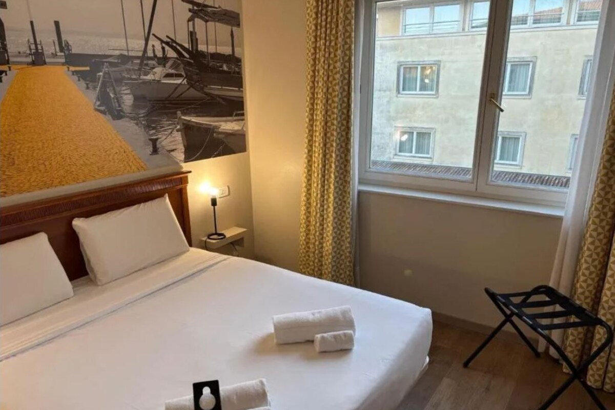 Une chambre d’hôtel avec un lit, une table de chevet, une lampe, une fenêtre avec des rideaux et une vue sur les bâtiments environnants.