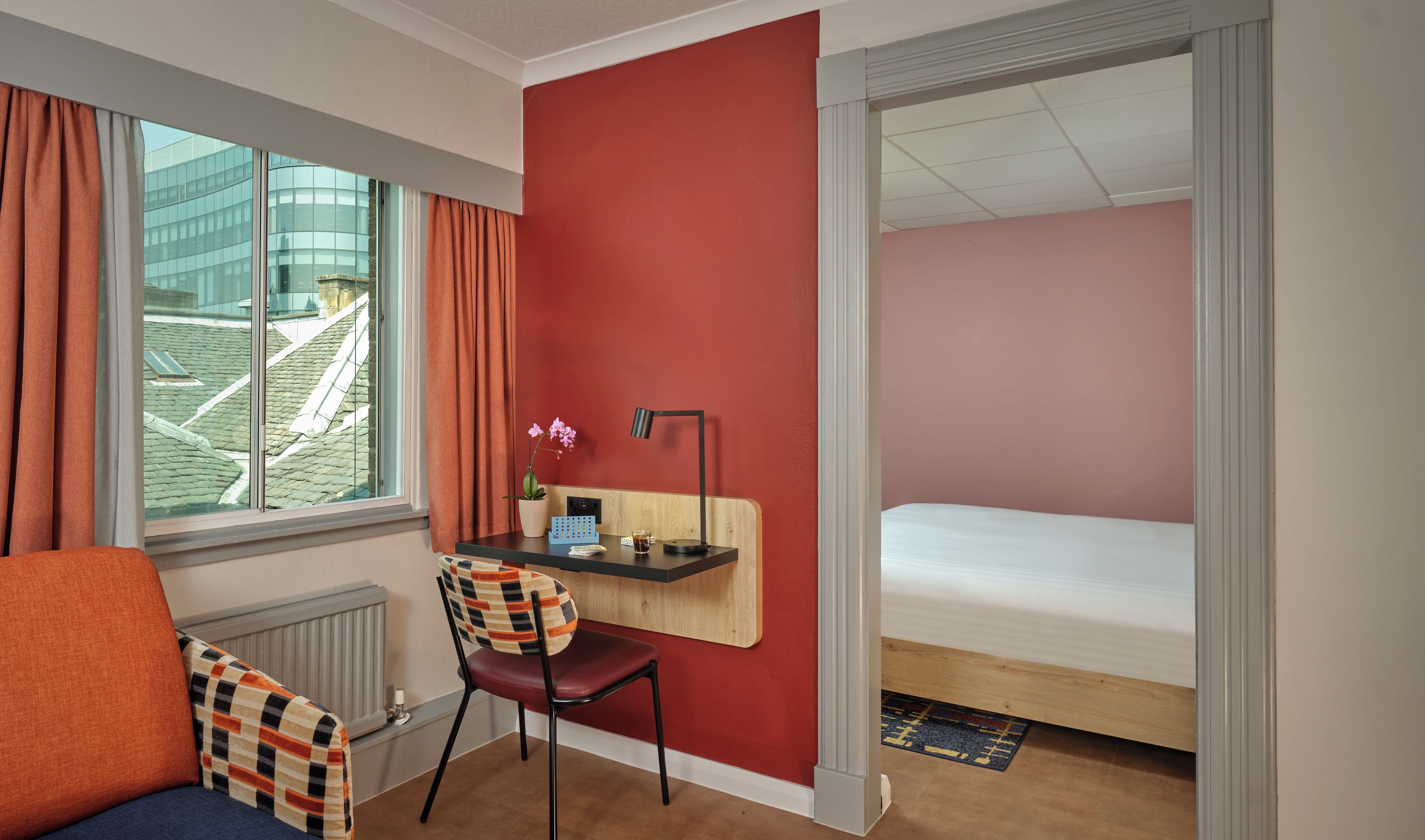 Een hotelkamer met een bed, een bureau met stoel, een raam met gordijnen en uitzicht op een gebouw.