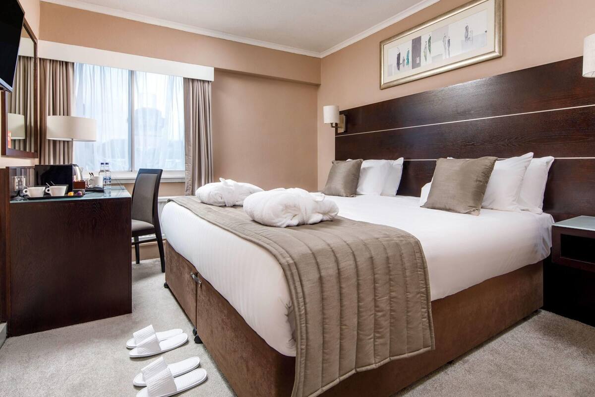 Une chambre d’hôtel avec un grand lit, un bureau et une chaise. Il y a une fenêtre avec des rideaux, un tableau encadré au mur et une paire de chaussons sur le sol.
