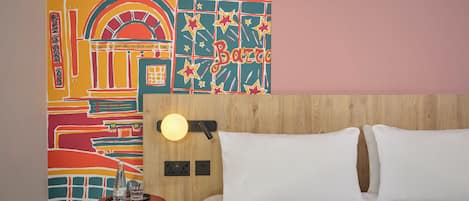 A hotel room with a colorful wall mural, a wooden headboard, a bedside table with a lamp, and a bed with white linens.