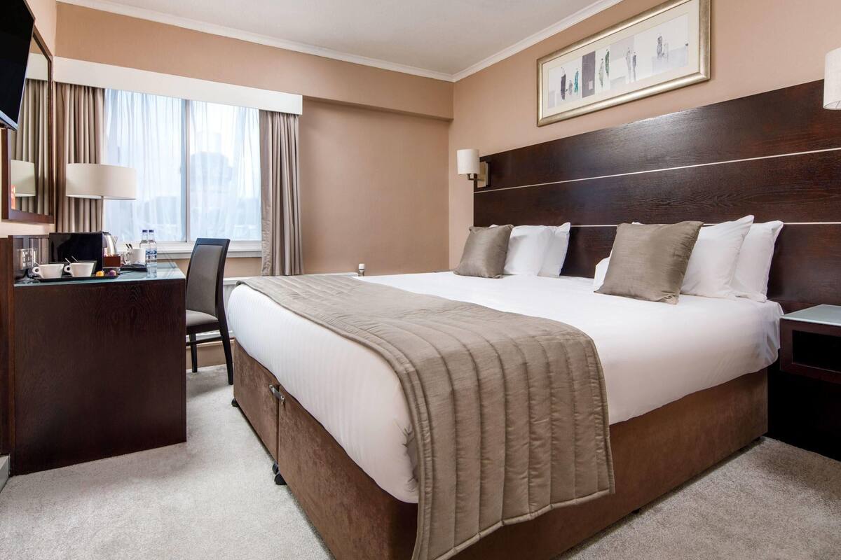 Une chambre d’hôtel avec un grand lit, un bureau, une chaise, une télévision et une fenêtre avec des rideaux.