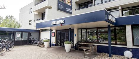Un edificio moderno con un letrero que dice "ENTREE". Hay bicicletas estacionadas afuera, un banco y una jardinera grande.