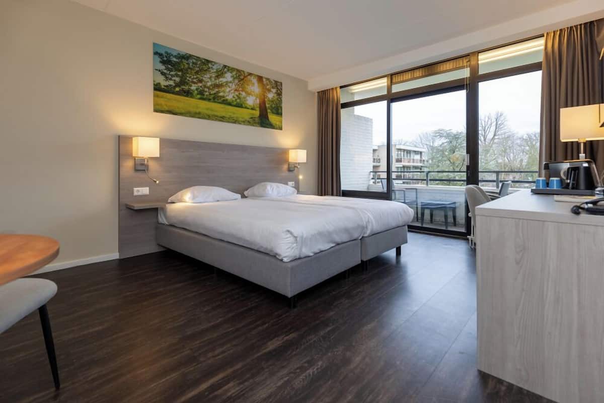 A hotel room with a bed, bedside tables, a desk, and a large window.
