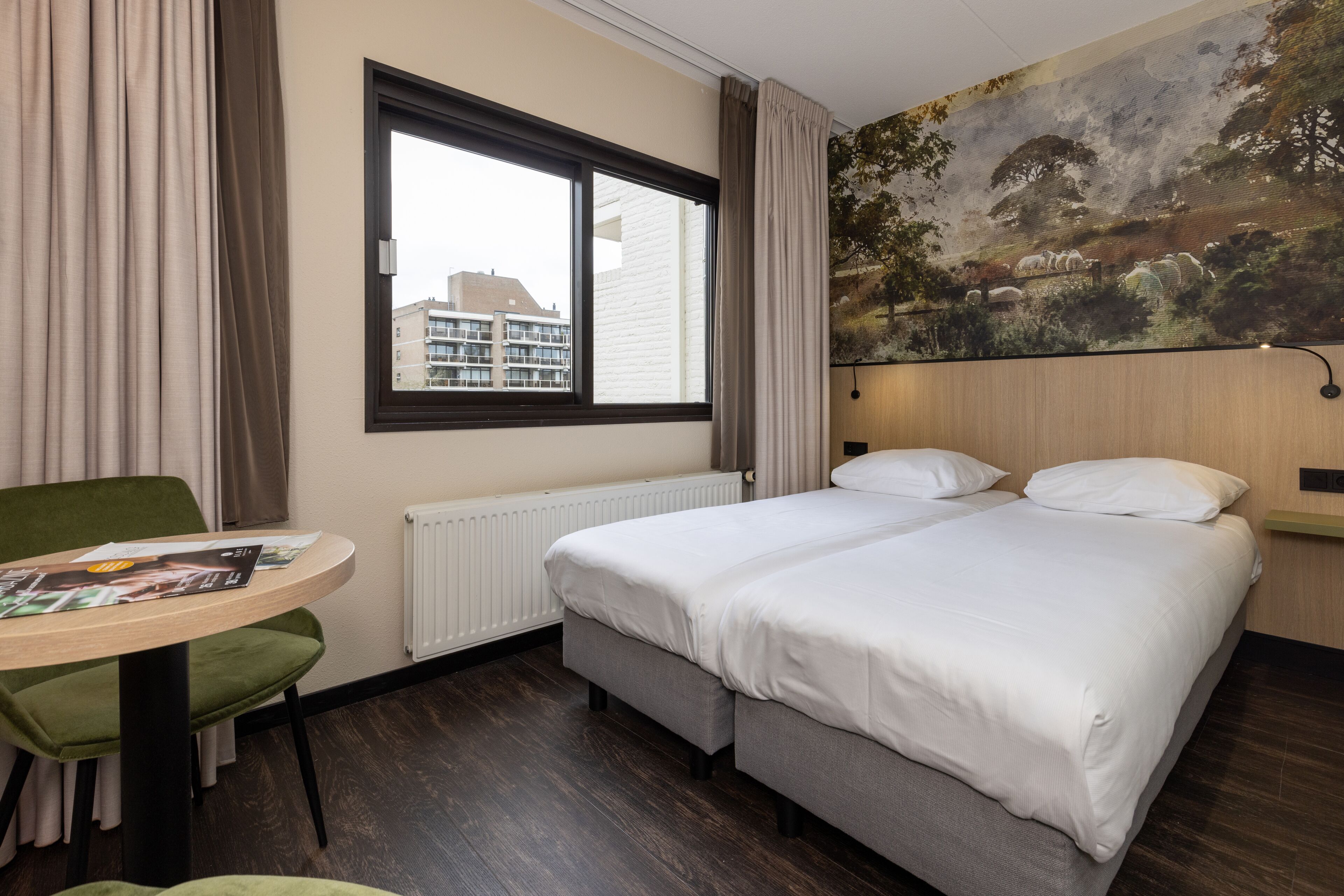 A hotel room with a bed, a table with a magazine, a green chair, a window, and a mural on the wall.