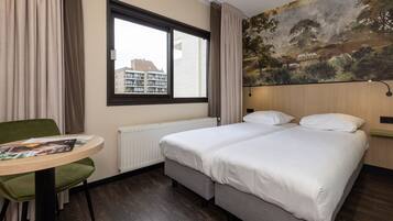 A hotel room with a bed, a table with a magazine, a green chair, a window, and a mural on the wall.