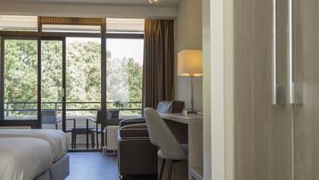Habitación de hotel con una cama, un escritorio, una silla y vistas a árboles a través de amplias ventanas.