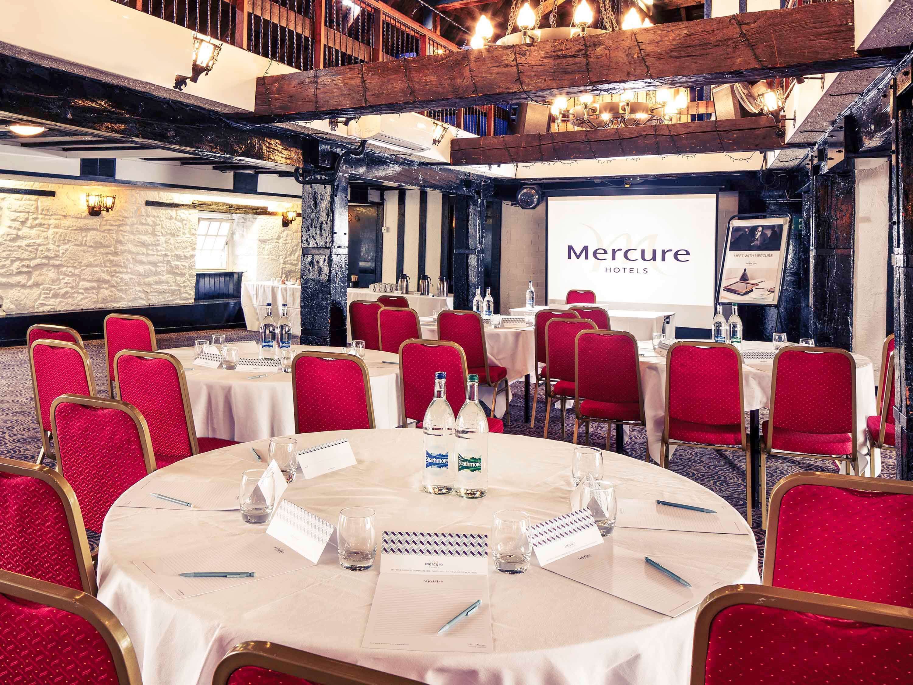 Una sala conferenze con tavoli rotondi, sedie rosse e un grande schermo che mostra 'Mercure Hotels'.