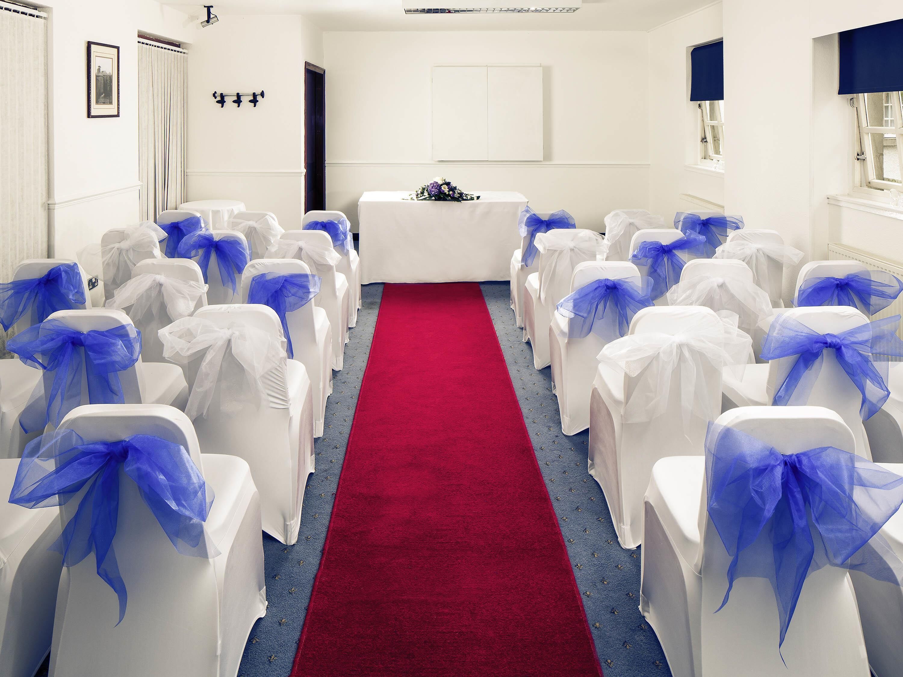 Um salão com fileiras de cadeiras brancas decoradas com laços azuis, um corredor com tapete vermelho e uma mesa branca com arranjo floral.