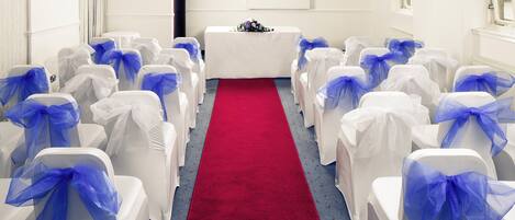Um salão com fileiras de cadeiras brancas decoradas com laços azuis, um corredor com tapete vermelho e uma mesa branca com arranjo floral.