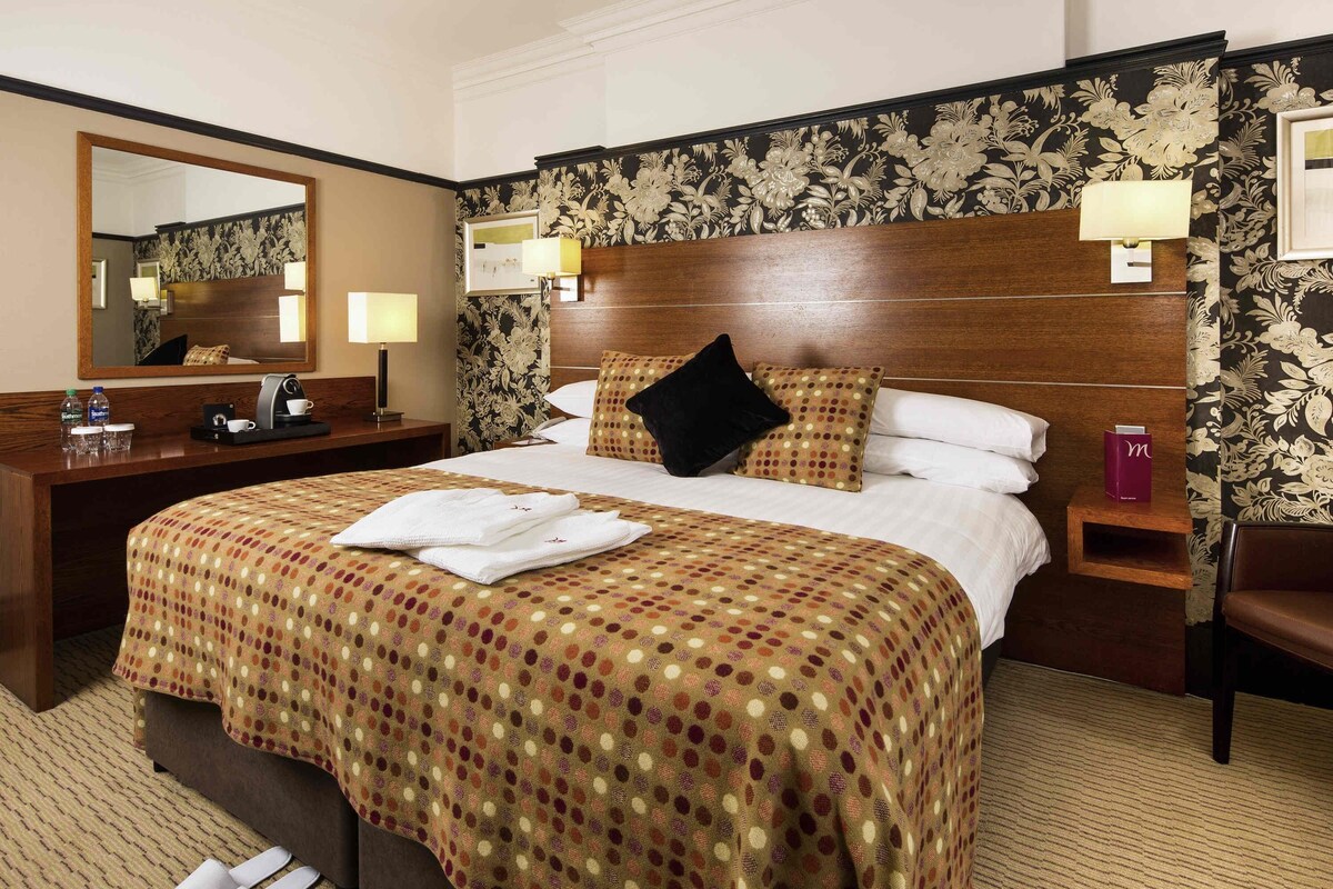 Habitación de hotel con una cama grande, cabecera de madera, mesita de noche con lámpara, escritorio con espejo y una silla.