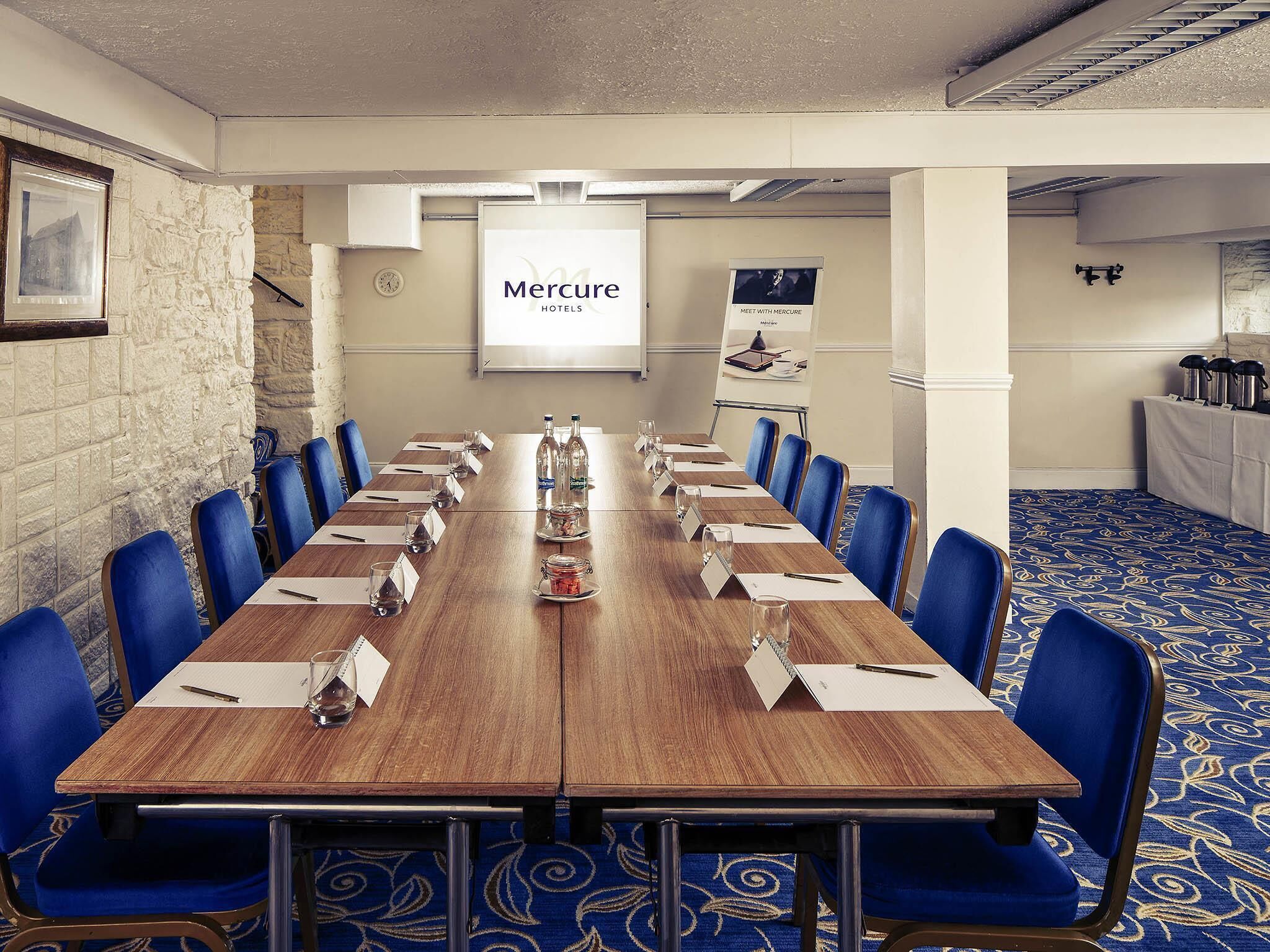 A conference room with a long table, blue chairs, nameplates, water bottles, and a 'Mercure Hotels' sign.