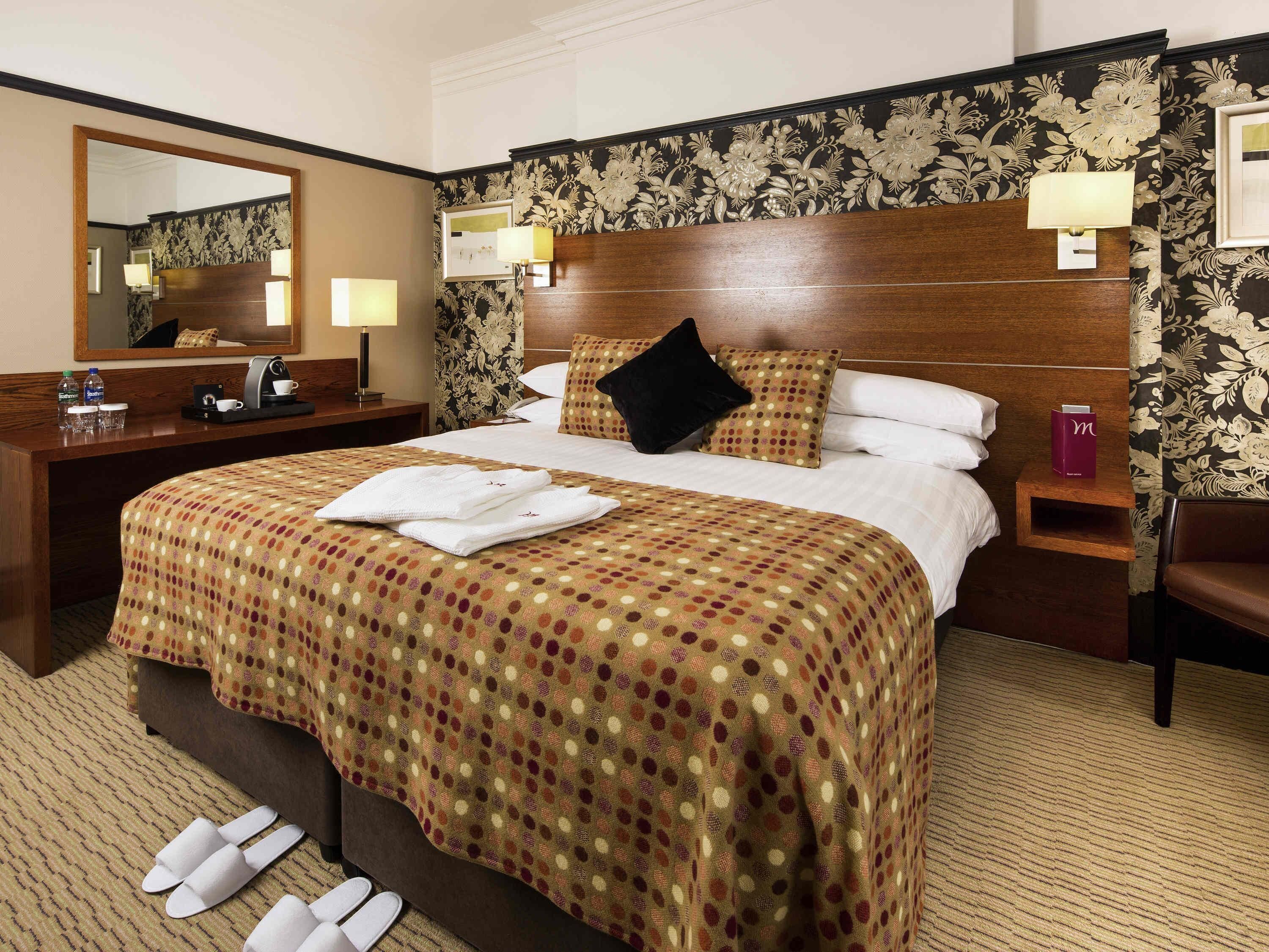 A hotel room with a large bed, a wooden headboard, a nightstand with a lamp, a desk with a mirror, and a chair.