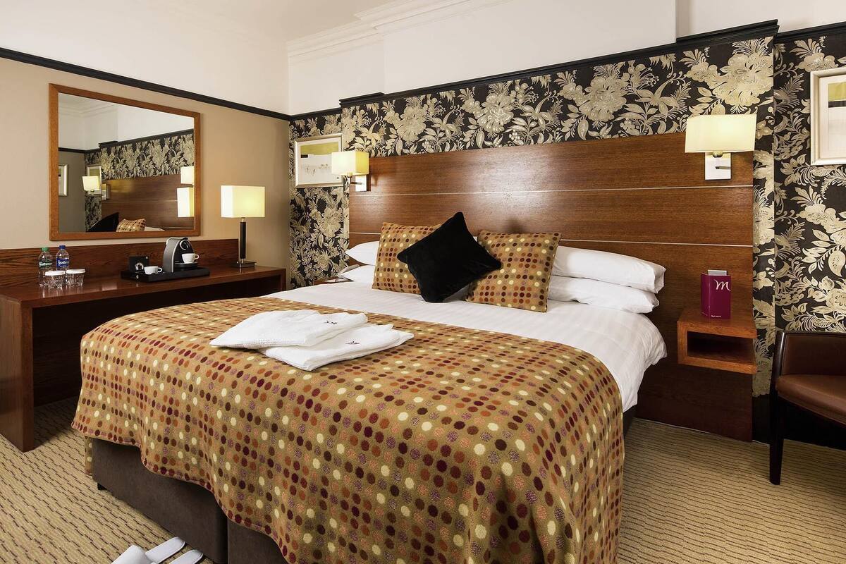 A hotel room with a large bed, a wooden headboard, a nightstand with a lamp, a desk with a mirror, and a chair.