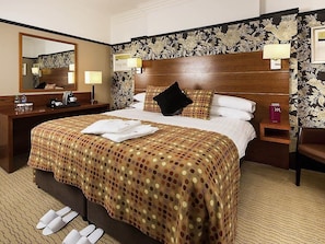 Habitación de hotel con una cama grande, cabecera de madera, mesita de noche, espejo y piso alfombrado.