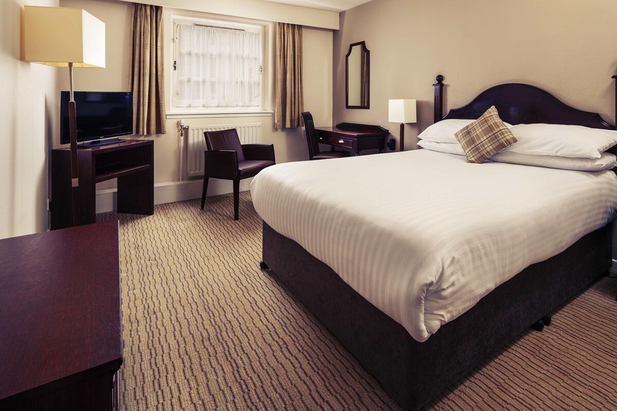 Habitación de hotel con una cama, un escritorio con televisión, una silla, una ventana con cortinas y un espejo en la pared.
