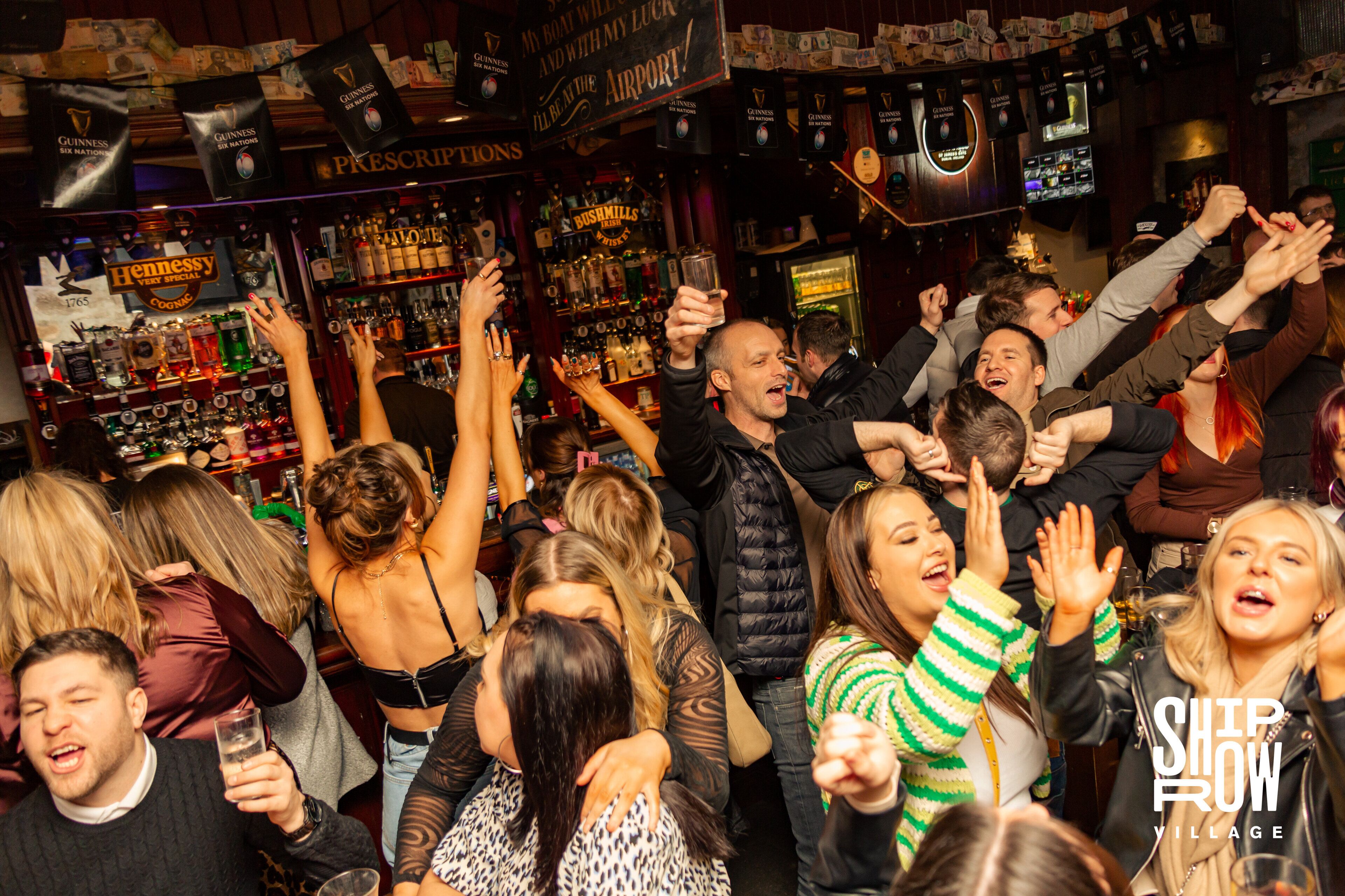 A crowded bar with people raising their hands, various liquor bottles on shelves, and a sign that reads "PRESCRIPTIONS".
