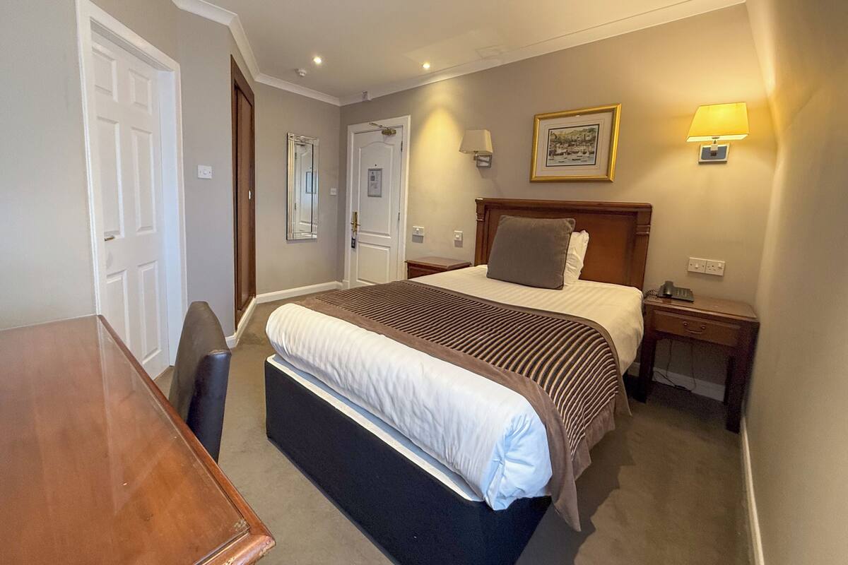 A hotel room with a bed, bedside table, lamp, and a framed picture on the wall.