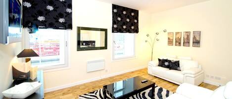 Una sala de estar moderna con un sofá blanco, alfombra estampada en blanco y negro, y una mesa de centro de vidrio. Hay tres ventanas con cortinas florales negras, un espejo y cuadros en la pared.