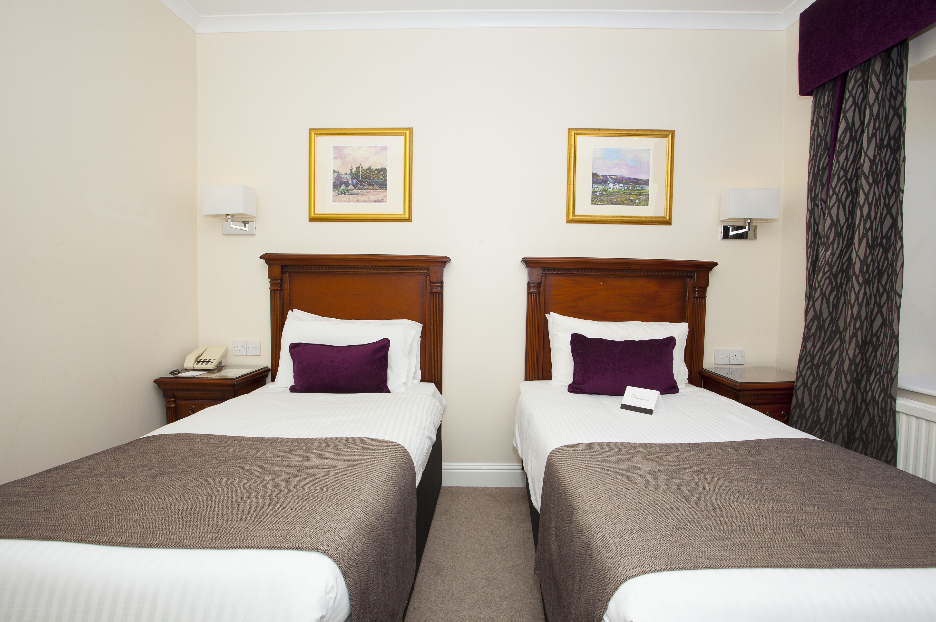 A hotel room with two single beds, a nightstand with a phone, and two framed pictures on the wall.