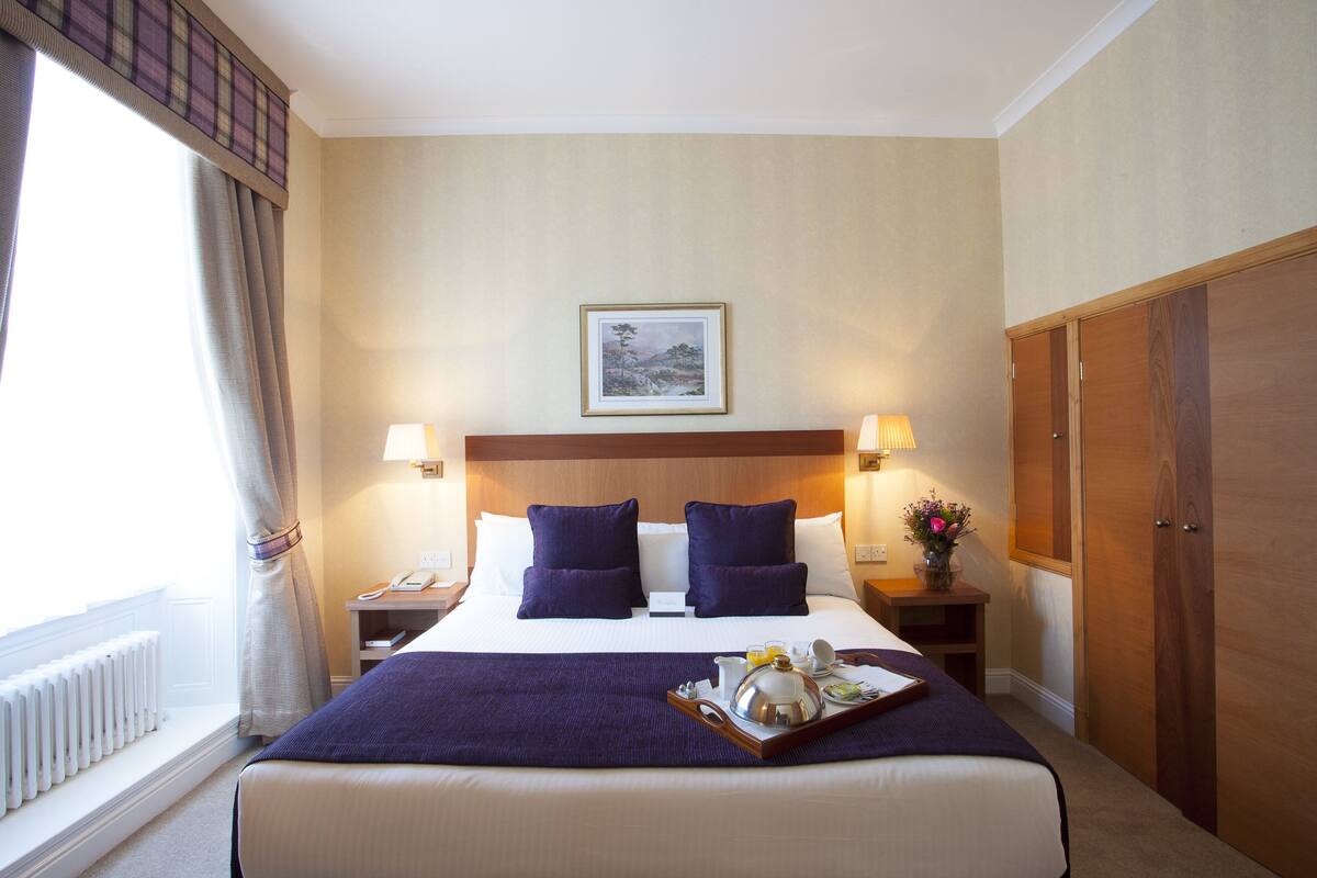 A hotel room with a large bed, two bedside tables, a nightstand, a wooden wardrobe, and a window with curtains.